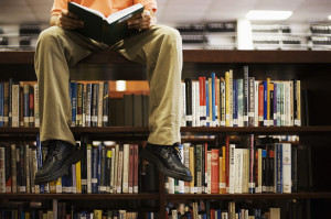 Man Reading Book and Sitting on Bookshelf in Library --- Image by © Royalty-Free/Corbis